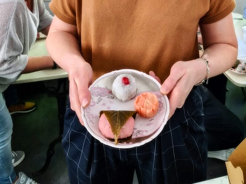 Personne tenant une assiette avec un mochi blanc, un gâteau rose enveloppé dans une feuille et une pâtisserie orange.