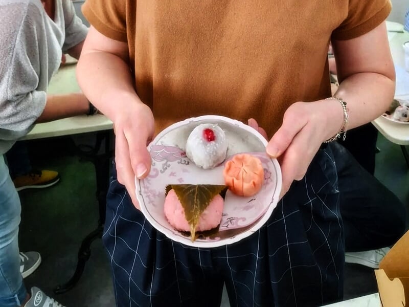 Personne tenant une assiette avec un mochi blanc, un gâteau rose enveloppé dans une feuille et une pâtisserie orange.