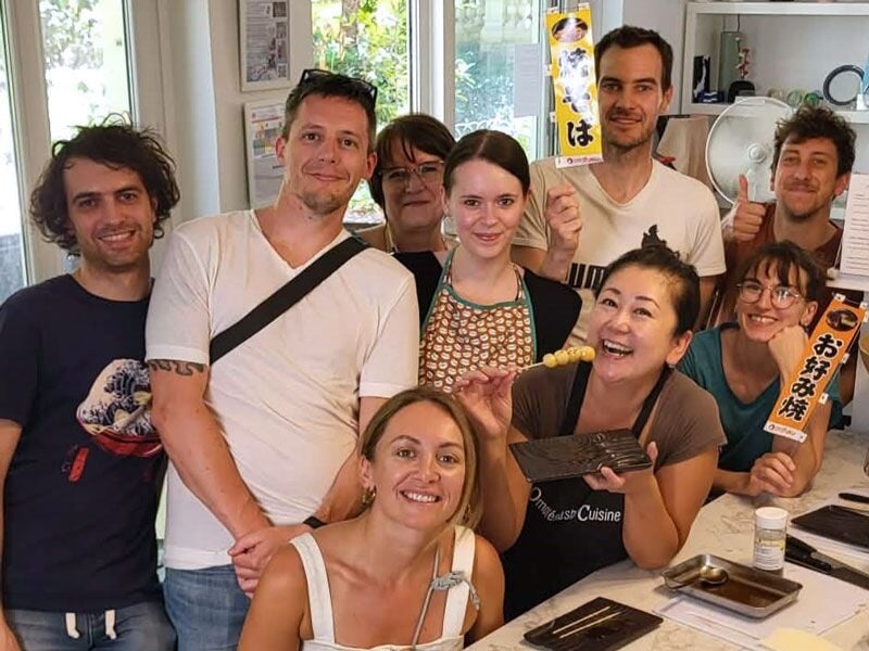 Groupe de huit personnes posant ensemble dans une cuisine, tenant des accessoires de cuisine japonaise, souriants.