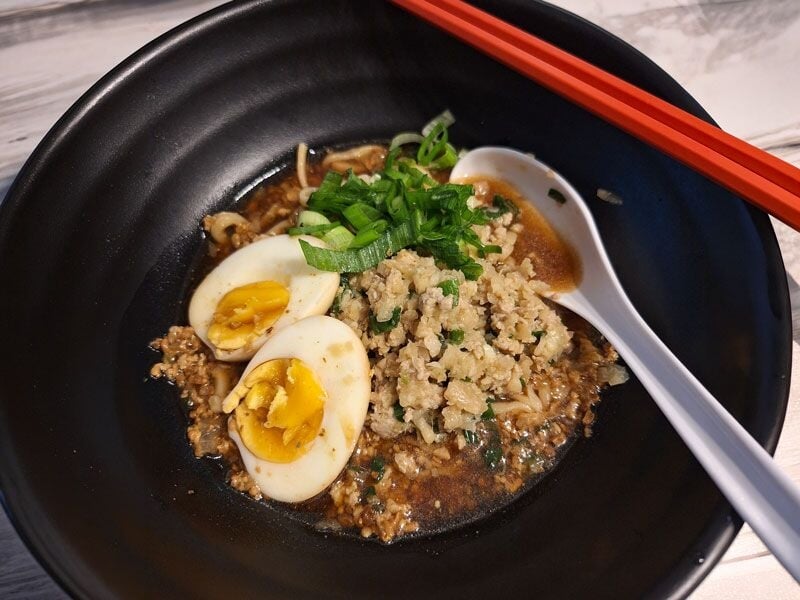 Bol noir contenant un plat asiatique avec œufs coupés, viande hachée, herbes fraîches et baguettes rouges.