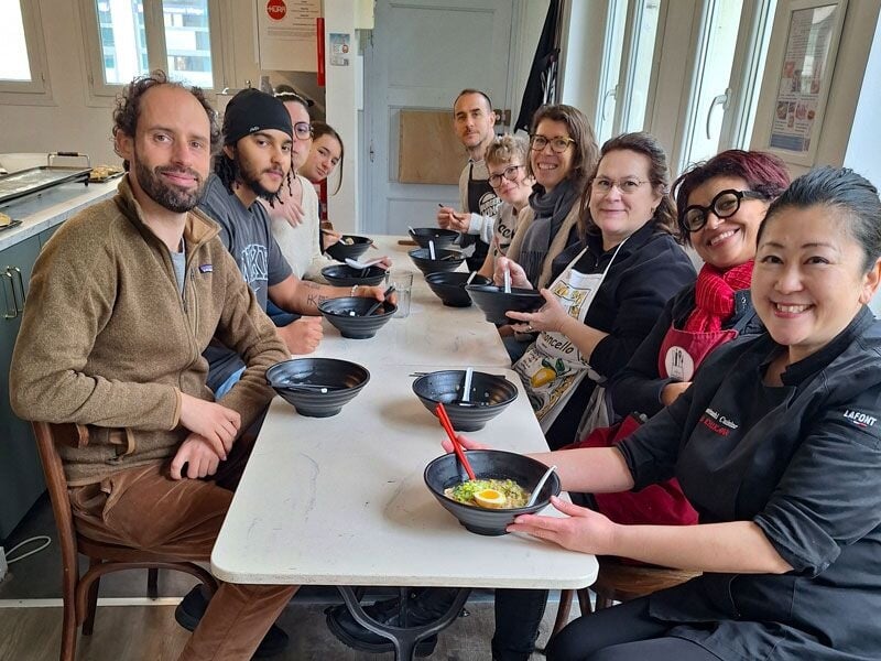 Groupe de personnes assises autour d'une table, dégustant un plat asiatique dans une ambiance chaleureuse et conviviale.