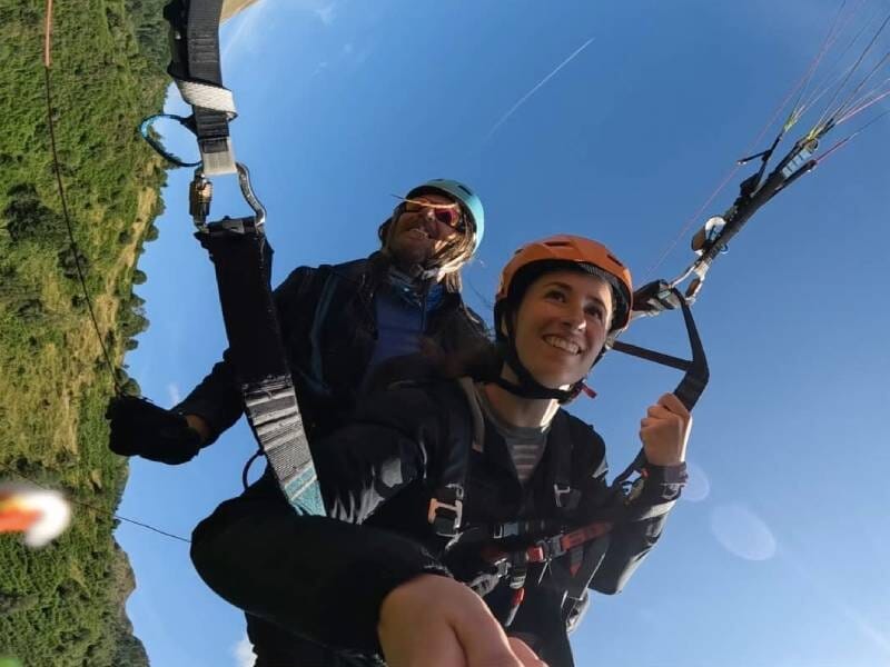 Deux personnes en parapente en tandem, souriantes, volant au-dessus d'un paysage vert sous un ciel bleu clair.
