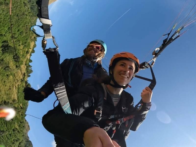 Deux personnes en parapente en tandem, souriantes, volant au-dessus d'un paysage vert sous un ciel bleu clair.