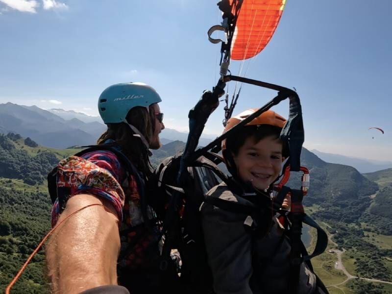 Un enfant et un adulte en parapente en tandem, survolant des montagnes verdoyantes sous un ciel dégagé.