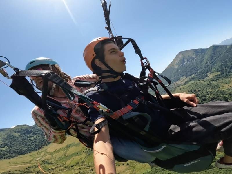 Deux personnes en parapente en tandem, survolant un paysage montagneux sous un ciel ensoleillé.