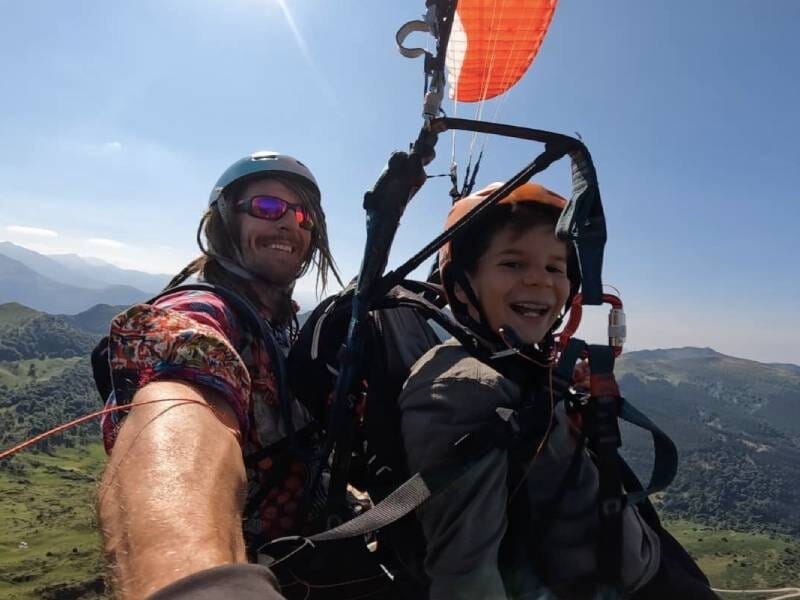 Deux personnes en parapente en tandem au-dessus des montagnes, souriantes et équipées de casques et harnais de sécurité lors d'un baptême près de Foix en Ariège.