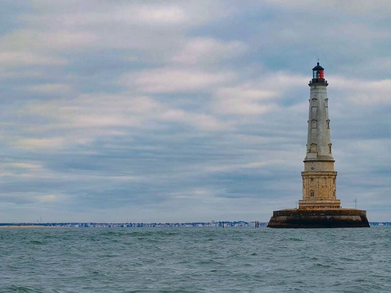 Phare blanc sur une base en pierre dans la mer sous un ciel nuageux, horizon avec une ville lointaine visible.