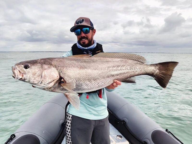 Un pêcheur souriant tient un gros poisson argenté sur un bateau gonflable en mer sous un ciel nuageux.