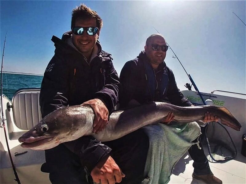 Deux hommes souriants sur un bateau tenant un énorme poisson serpenté lors d'une sortie de pêche en mer.