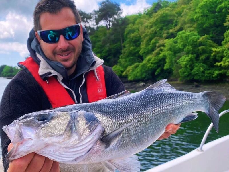 Homme avec lunettes de soleil tenant un gros poisson argenté sur un bateau, avec un paysage vert en arrière-plan.
