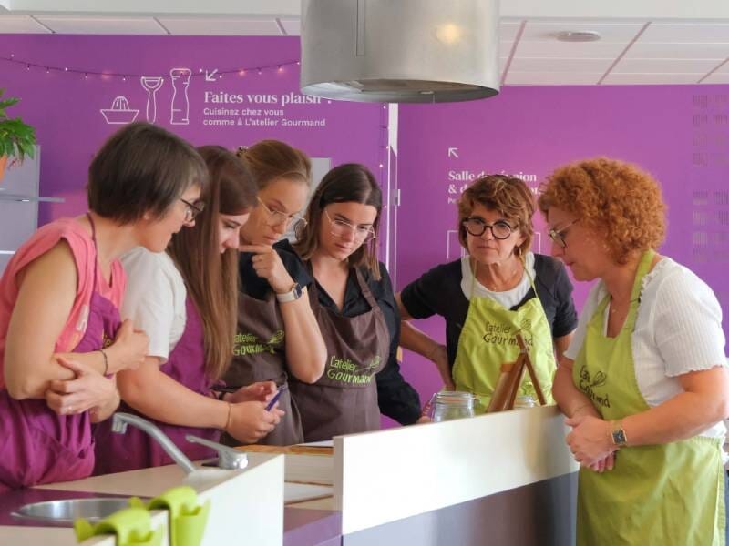 Groupe de cinq femmes concentrées lors d'un cours de cuisine dans un atelier avec tabliers verts et mur violet.