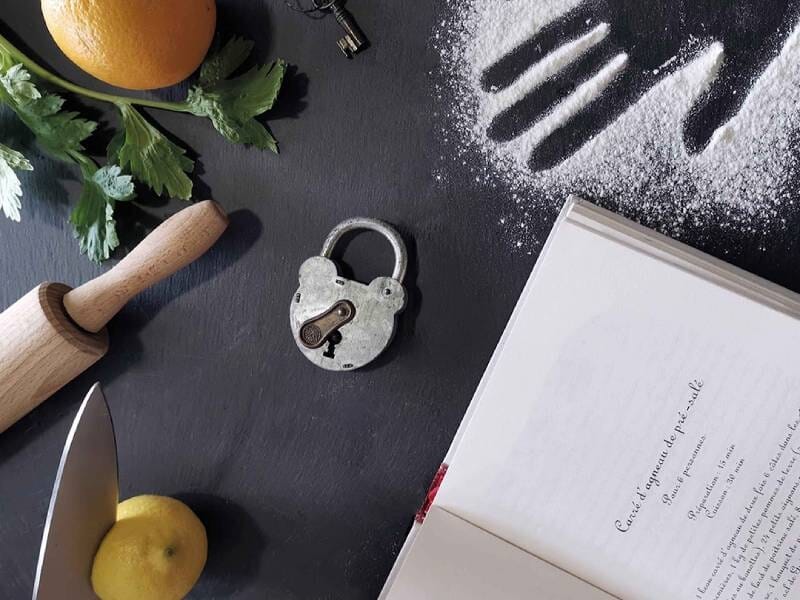 Plan de travail avec rouleau à pâtisserie, citron, cadenas ancien, traces de farine et un livre de recettes ouvert.
