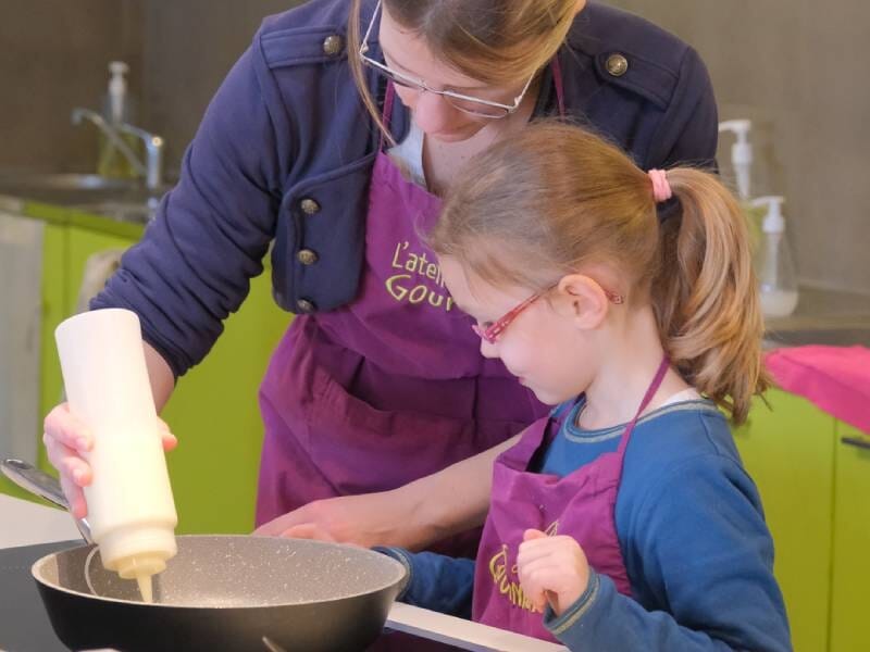 Une femme aide une petite fille à verser un liquide dans une poêle lors d'un cours de cuisine en atelier près de Vannes.
