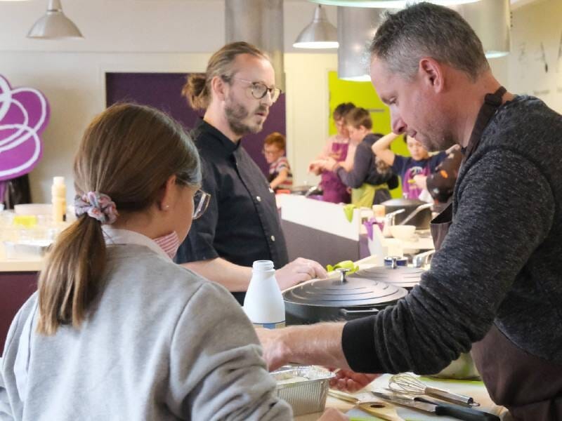 Des adultes et enfants participent à un cours de cuisine dans une salle équipée avec des plans de travail.