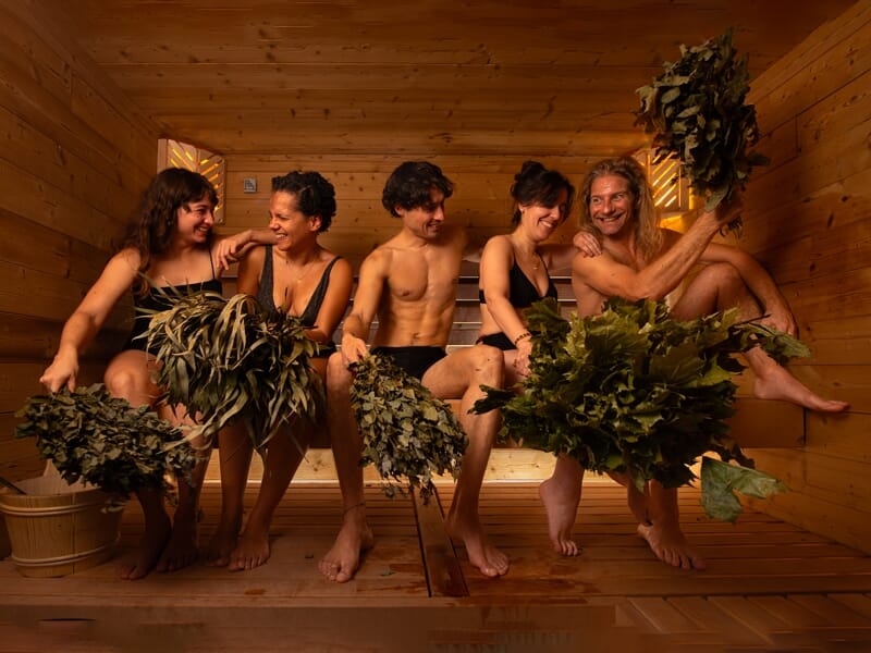 Quatre personnes assises dans un sauna en bois, tenant des bouquets de feuilles pour un rituel de bien-être traditionnel.