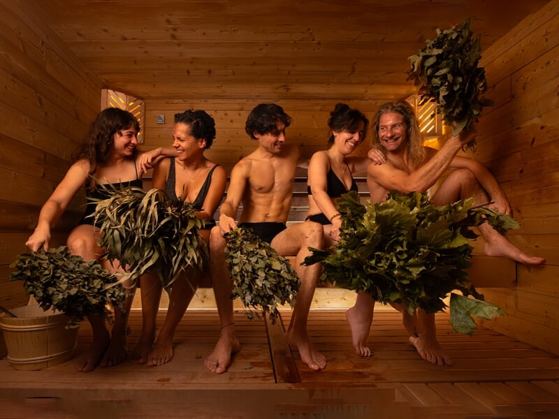 Quatre personnes assises dans un sauna en bois, tenant des bouquets de feuilles pour un rituel de bien-être traditionnel.