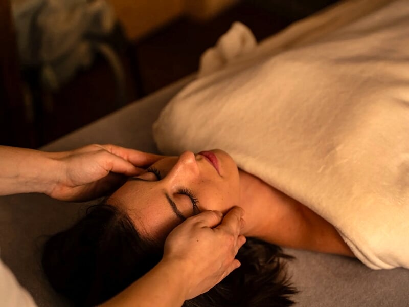 Une femme reçoit un massage du visage allongée sur une table, enveloppée dans une couverture claire.