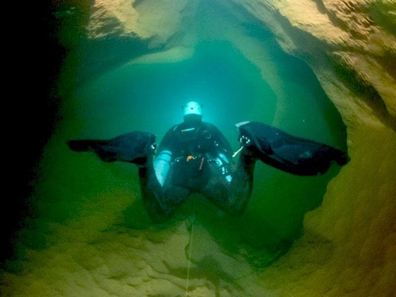 Plongeur équipé de palmes et lampe explorant une grotte sous-marine aux parois rocheuses jaunes.