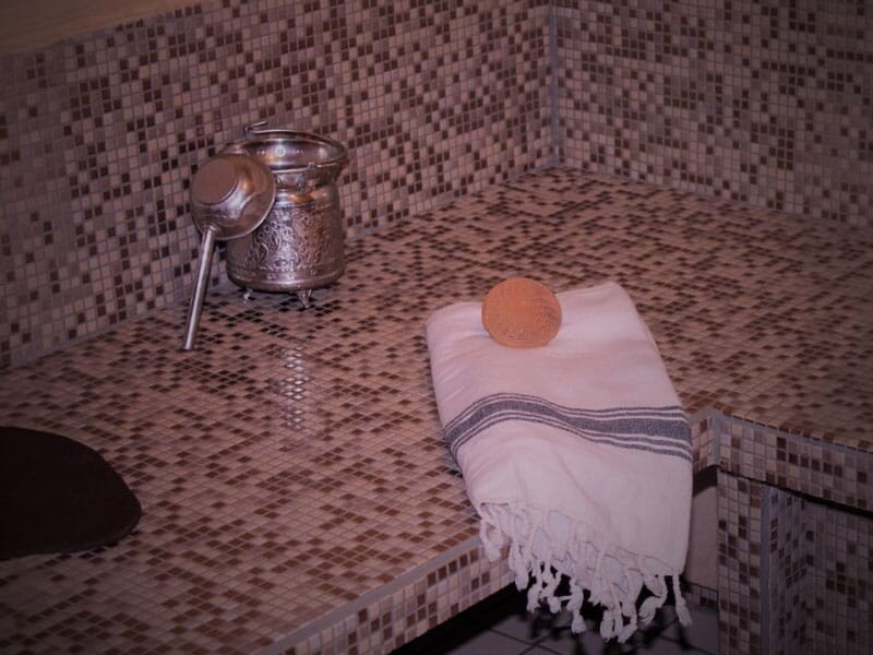 Un banc en mosaïque dans un hammam avec une serviette blanche, un savon rond et un récipient métallique posé dessus.