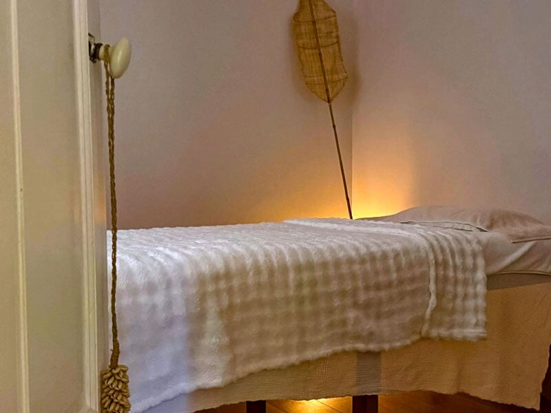 Table de massage avec couverture blanche dans une pièce calme éclairée par une lumière douce et chaleureuse.