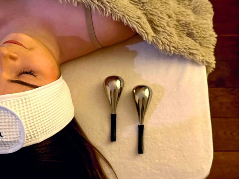 Femme allongée sur une table de massage avec bandeau blanc et deux outils de massage métalliques à côté.