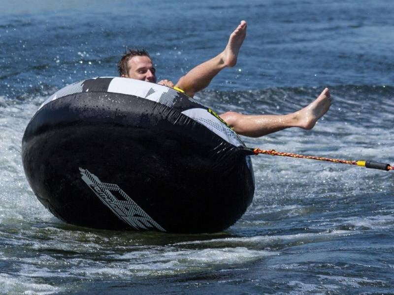 Un homme en combinaison noire est assis sur une bouée ronde noire tractée sur l'eau, les pieds levés en l'air.