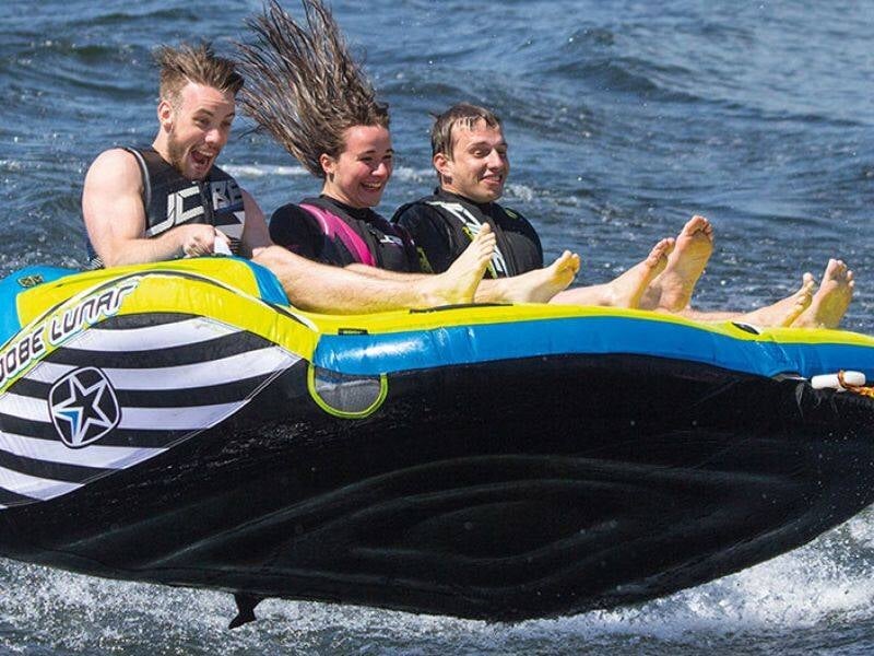 Trois personnes souriantes sur une grande bouée gonflable noire, jaune et bleue, tractée à grande vitesse sur l'eau, lors d'une session de bouée tractée à Pornic.