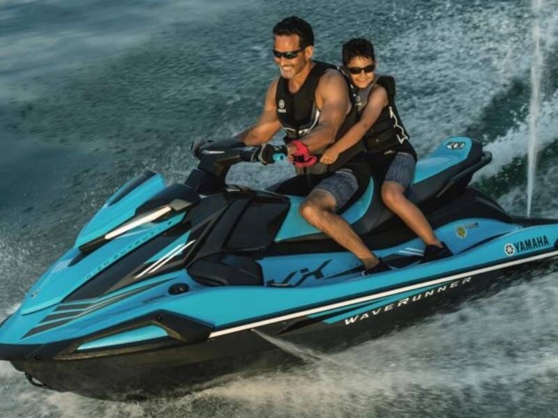 Deux personnes en gilets de sauvetage noirs sur un jet ski bleu Yamaha naviguant rapidement sur l'eau.