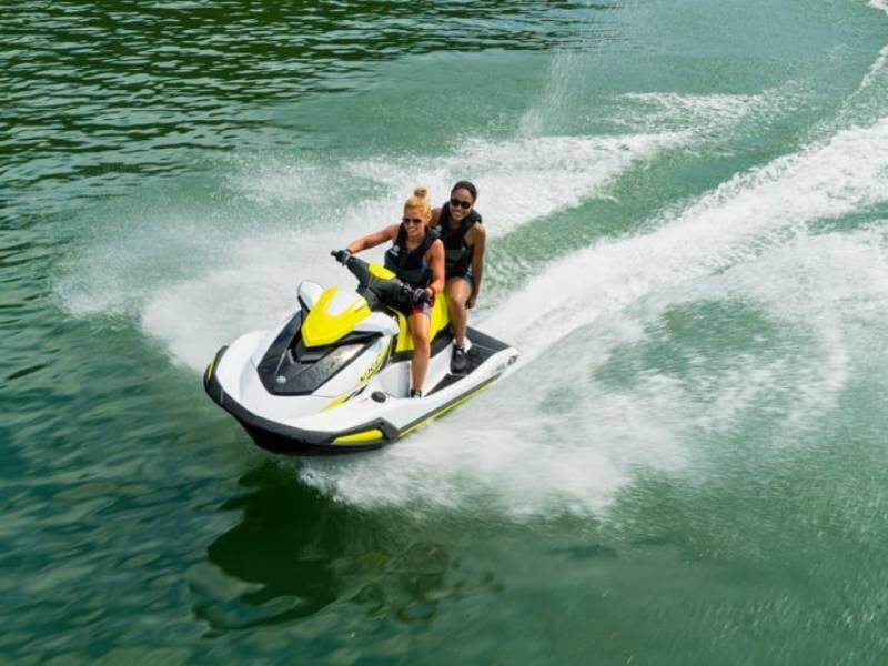 Deux femmes en gilets de sauvetage noirs sur un jet ski blanc et jaune glissant sur une eau verte calme.