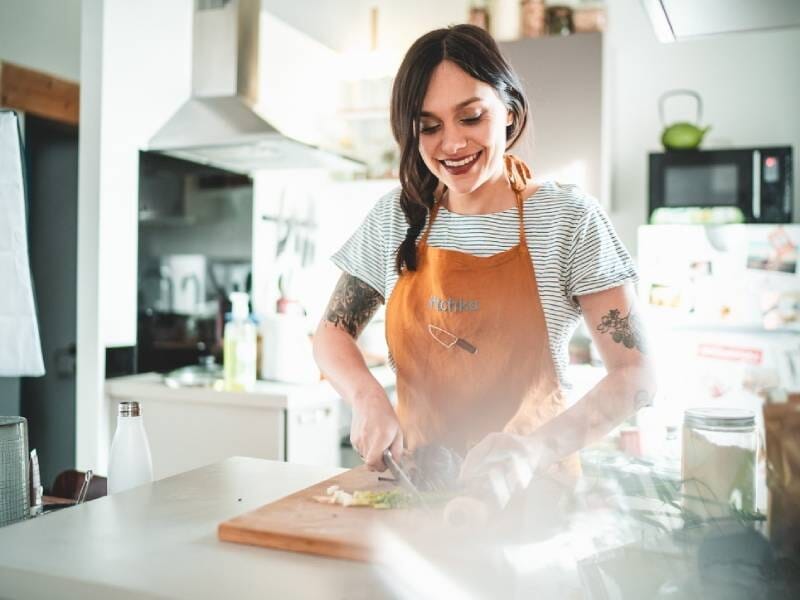 Jeune femme souriante en tablier orange préparant des légumes frais dans une cuisine moderne et lumineuse.