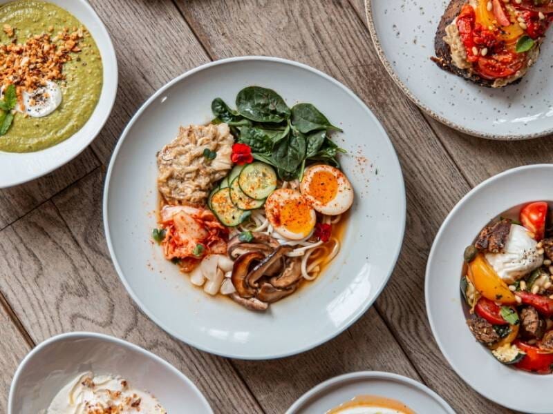 Assiette gourmande avec œufs, légumes variés et épinards, entourée d'autres plats colorés sur une table en bois.