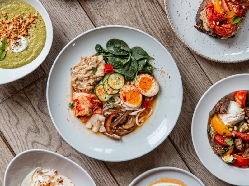 Assiette gourmande avec œufs, légumes variés et épinards, entourée d'autres plats colorés sur une table en bois.