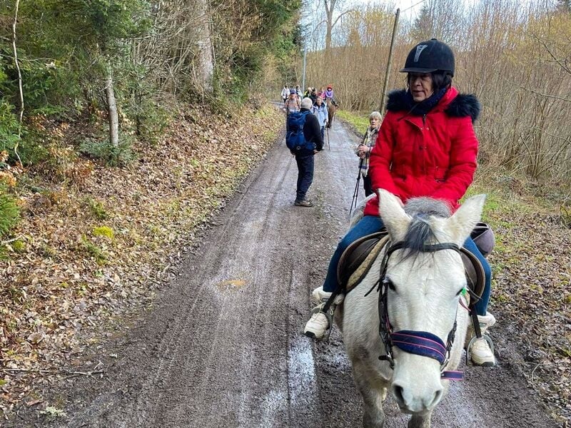 Femme en manteau rouge montant un cheval blanc sur un chemin boueux avec d'autres randonneurs à pied.