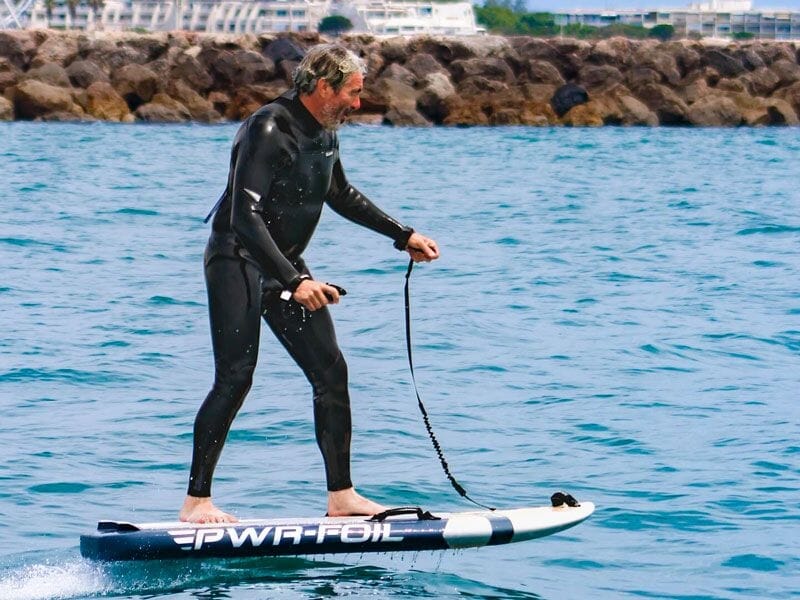 Homme en combinaison de plongée noire sur un surf électrique PWR-FOIL glissant sur l'eau près d'une digue rocheuse.