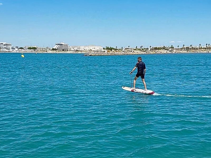 Homme en combinaison noire pratiquant le surf électrique sur une mer calme avec la ville en arrière-plan sous un ciel bleu.