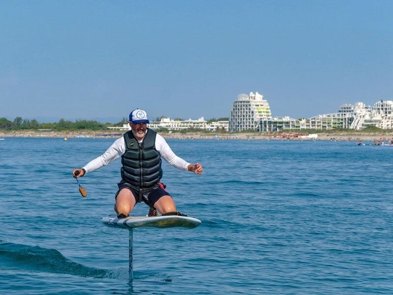 Homme souriant en gilet de sauvetage fait du surf électrique sur un foil au large de La Grande Motte par temps clair.