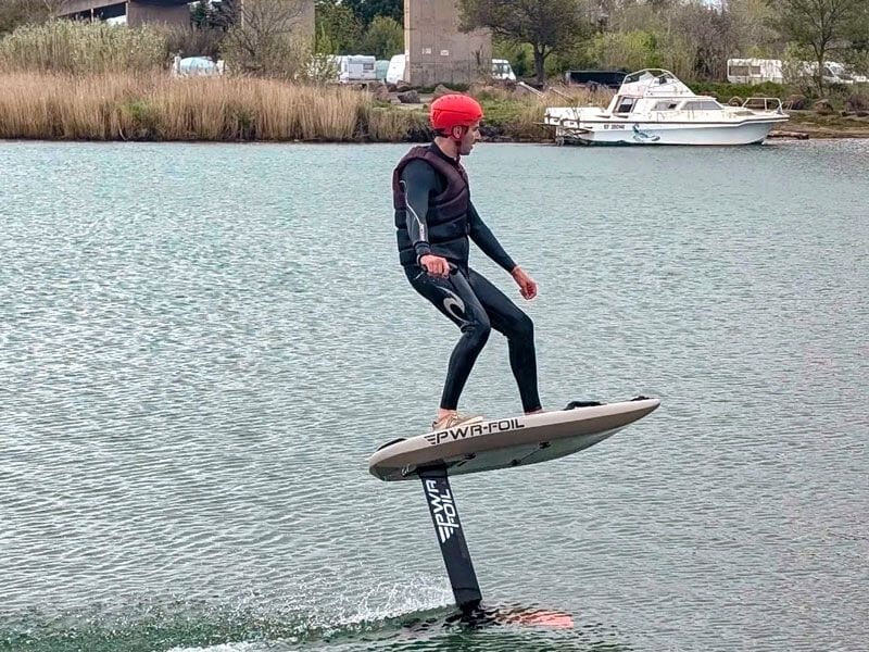 Homme en combinaison et casque rouge pratiquant le wingfoil sur un plan d'eau calme près d'une rive avec bateau.