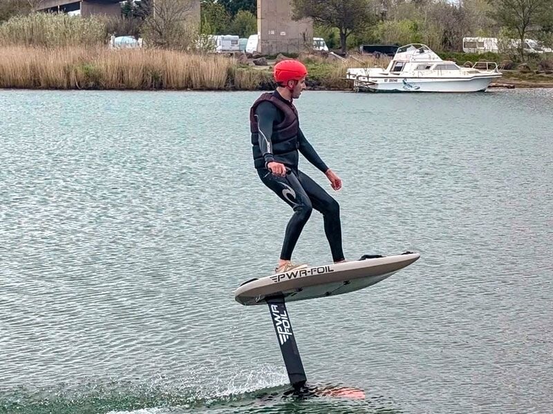 Homme en combinaison et casque rouge pratiquant le wingfoil sur un plan d'eau calme près d'une rive avec bateau.