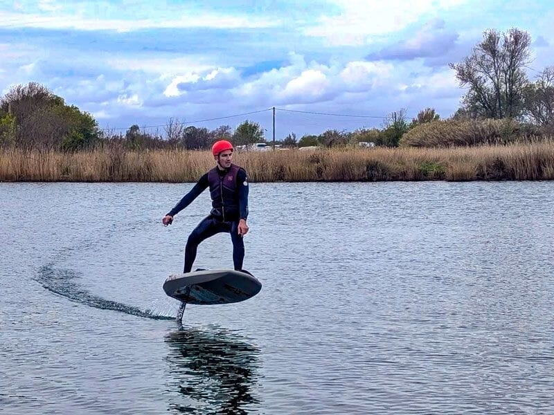 Personne en combinaison noire et casque rouge glissant sur une planche de wingfoil sur une eau tranquille avec végétation en arrière.