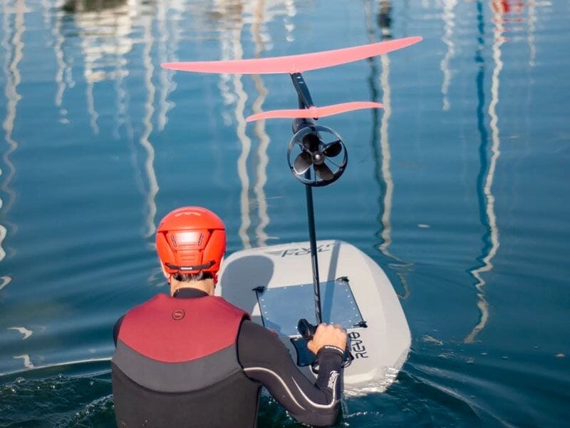 Vue arrière d'un pratiquant en combinaison et casque rouge tenant une planche de wingfoil avec moteur électrique sur l'eau.