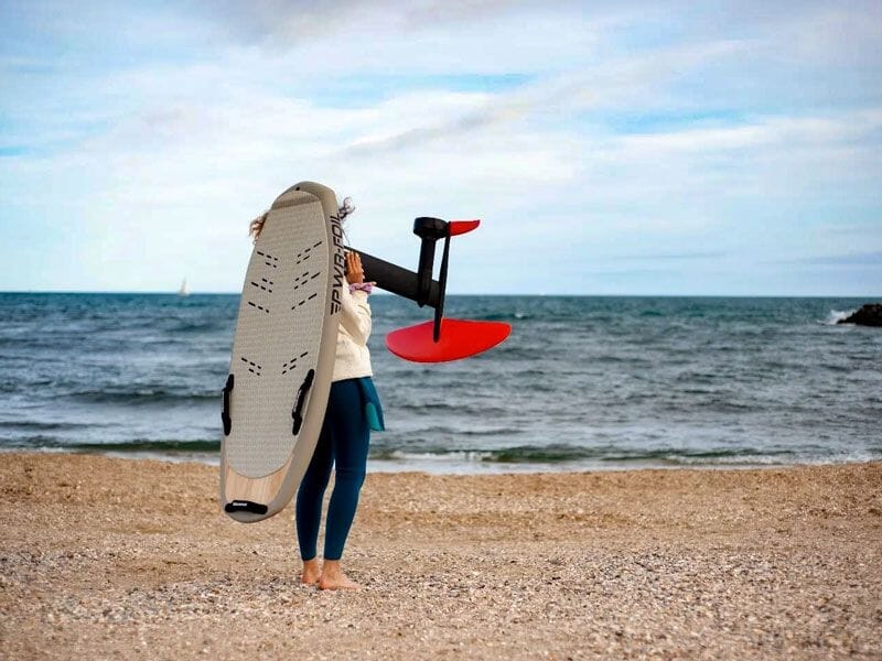 Femme debout sur une plage tenant une planche de wingfoil avec foil rouge, mer et ciel nuageux en arrière-plan.