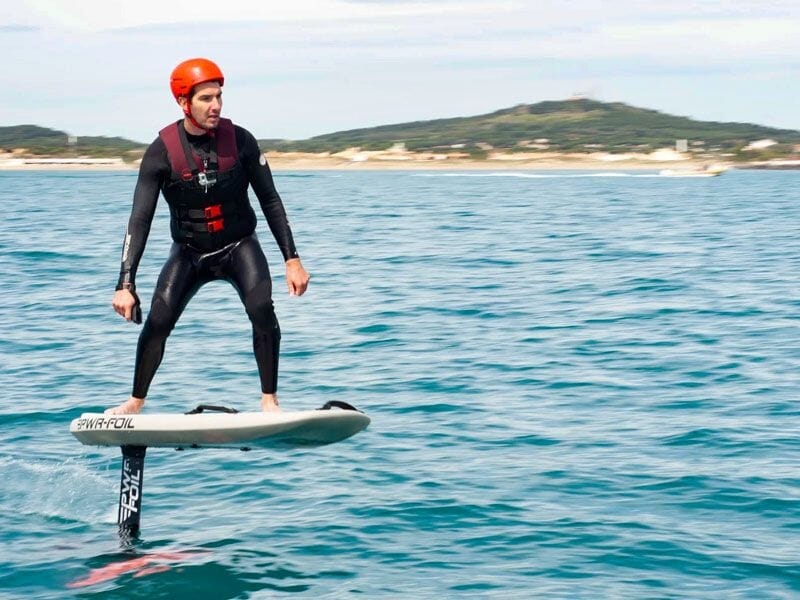 Homme en combinaison et casque rouge debout sur une planche de wingfoil en mer avec une colline en arrière-plan.