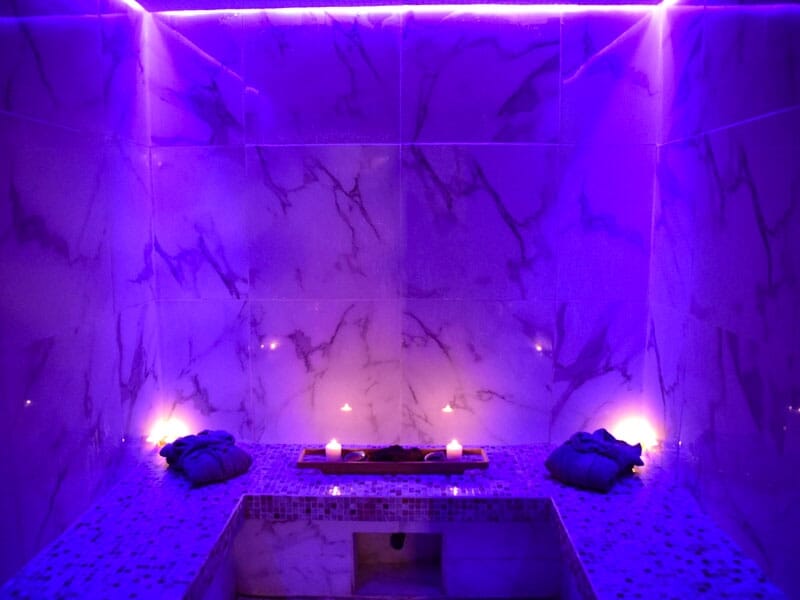 Intérieur d'un hammam avec murs en marbre, lumière violette et bougies pour une ambiance apaisante.