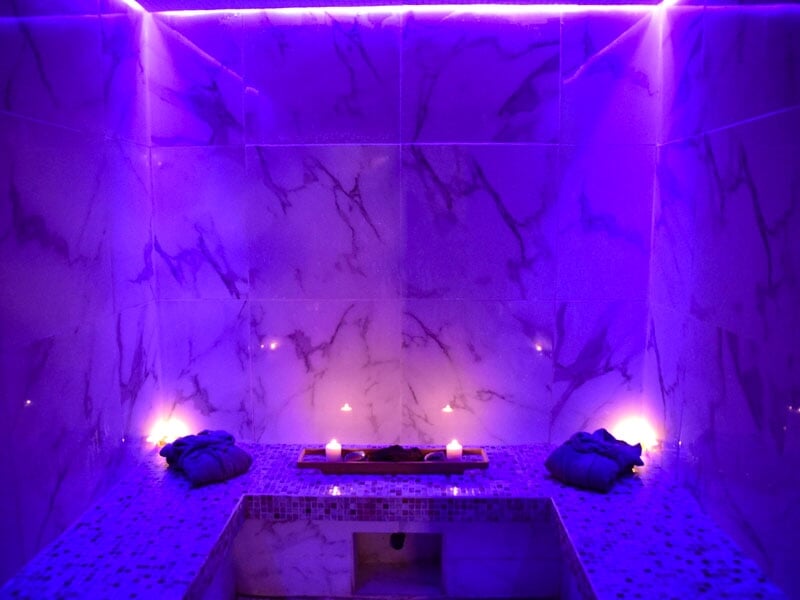 Intérieur d'un hammam avec murs en marbre, lumière violette et bougies pour une ambiance apaisante.
