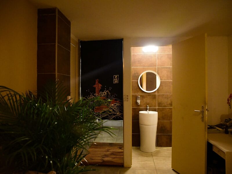Entrée d'une salle de spa avec plantes, miroir rond et lumière douce, ambiance chaleureuse.