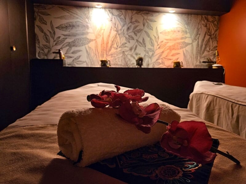Table de massage avec serviette roulée et orchidée rouge, ambiance tamisée et décor floral.