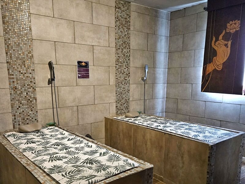 Salle de spa avec deux tables de massage en pierre, murs carrelés et lumière naturelle.