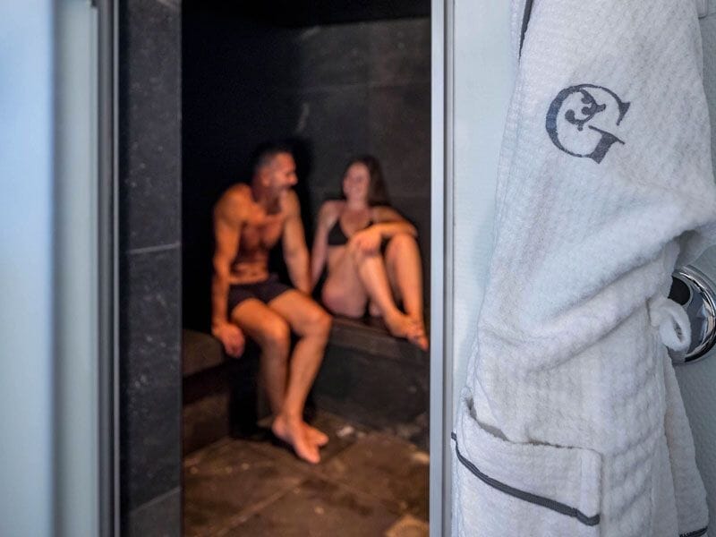 Deux personnes en maillot de bain se détendent dans un sauna sombre, avec un peignoir blanc suspendu à côté.