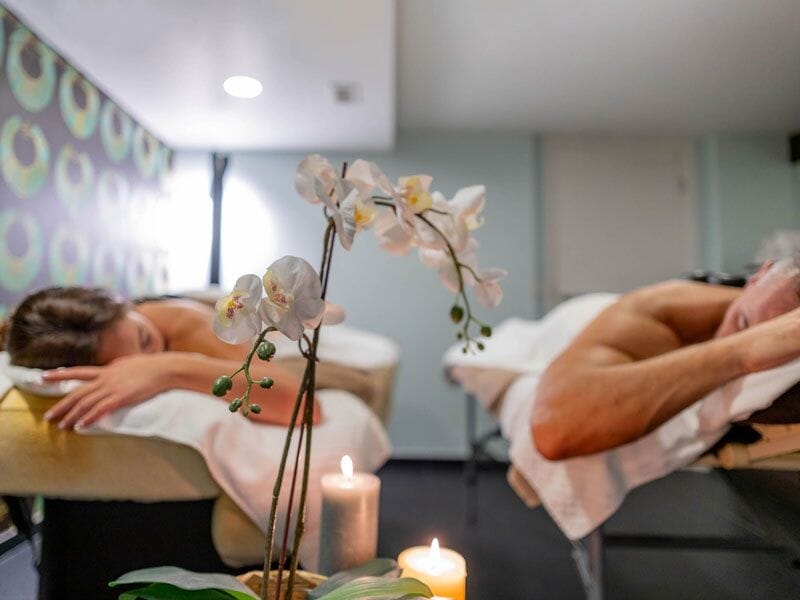Un couple allongé sur des tables de massage dans une salle de spa décorée avec des orchidées et une bougie.