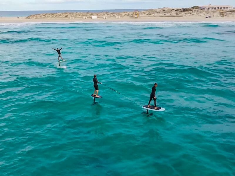 Trois personnes pratiquent le surf électrique sur une mer calme avec une plage et des dunes en arrière-plan.